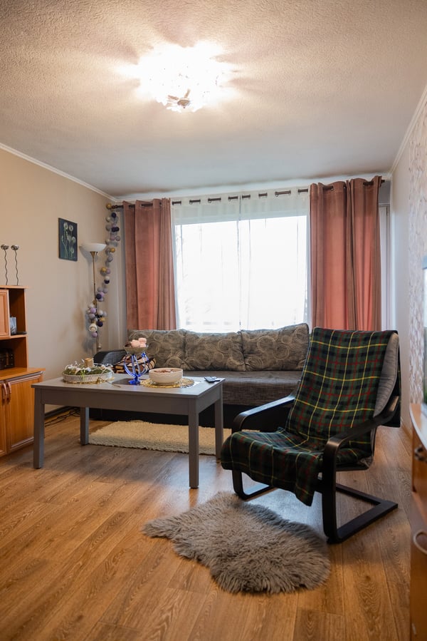 Гостиная с деревянным полом: у окна со шторами стоит диван, рядом журнальный столик, на переднем плане — кресло. Пространство практичное, но обставлено разностилевой мебелью.