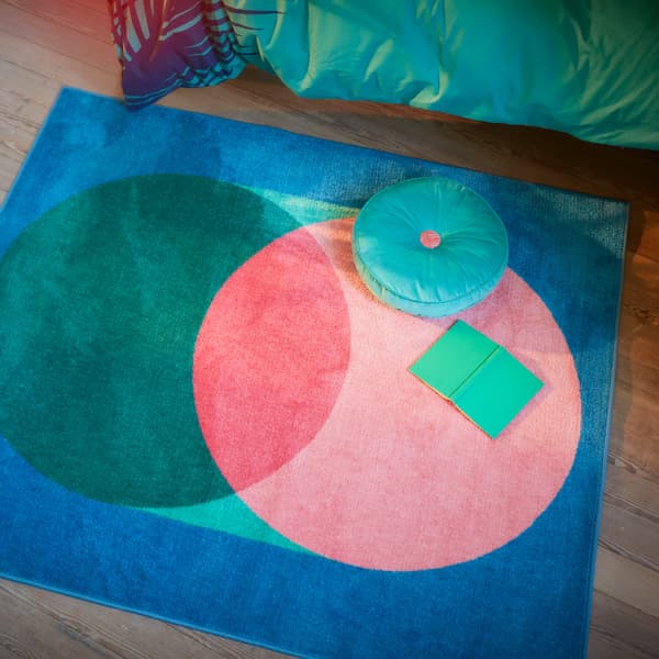 GRACIÖS rug with a big circular pattern in pink and green on a blue background, with a book and cushion placed on it.