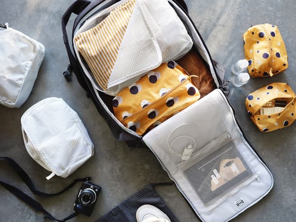 Grand sac de rangement de voyage ouvert par terre avec différents compartiments, des trousses de voyage à l'intérieur, un chargeur de téléphone et des documents à l'interieur de la pche de la fermeture. D'autres compartiments fermés sur le sol à côté du sac avec d'autres trousses.