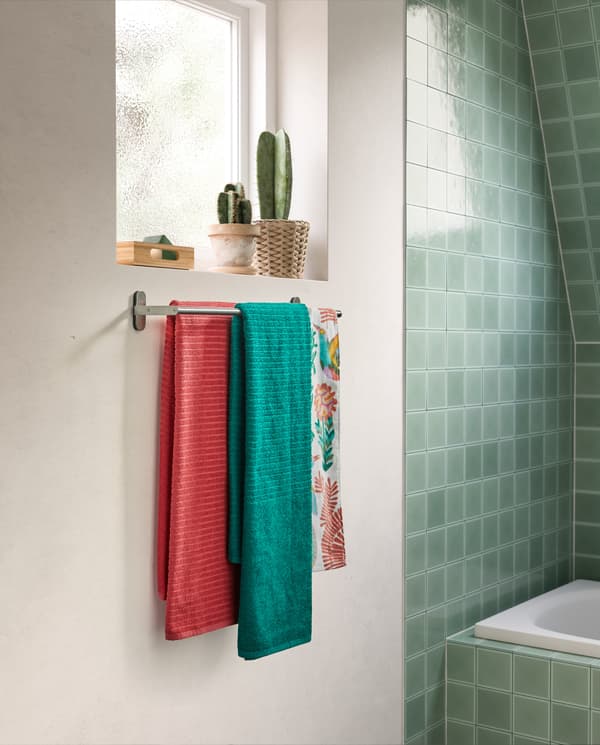 Green and pink towels with woven stripes hang on a BROGRUND towel rail in stainless steel that’s wall-mounted under a window.