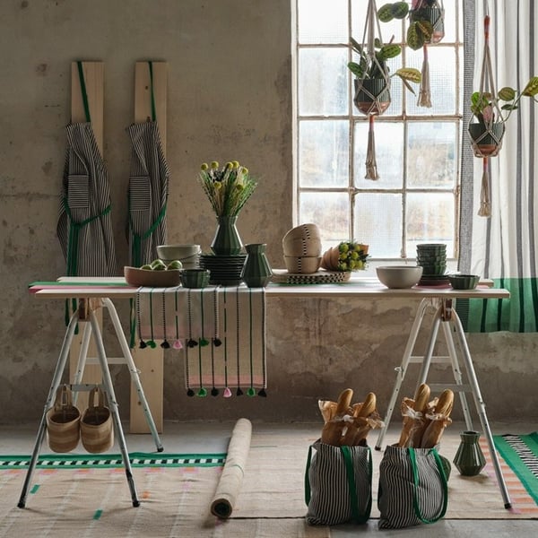 Green plates, bowls, and vases from the MÄVINN collection sit on a table in an industrial-style room. Two beige-green rugs lie beneath, and three plant pot hangers hang in the window.