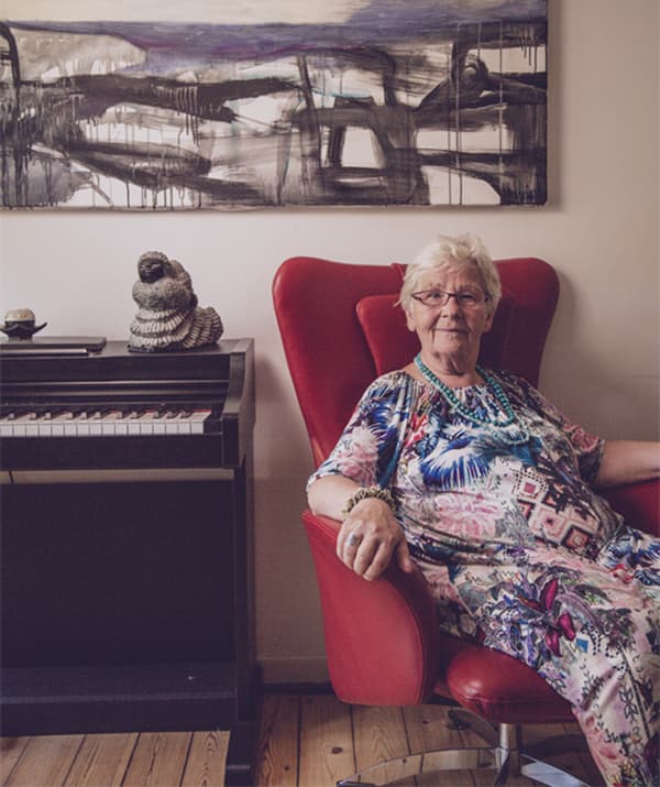 Greta นั่งอยู่บนเก้าอี้นวมสีแดงมีปีกถัดจากเปียโน ผนังด้านหลังมีงานศิลปะติดอยู่