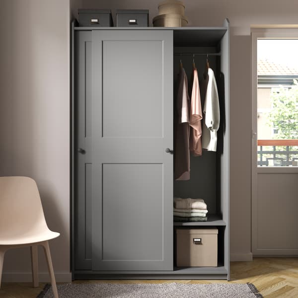 Grey HAUGA sliding-door wardrobe placed next to a door stands partly open, revealing storage boxes and garments on hangers.