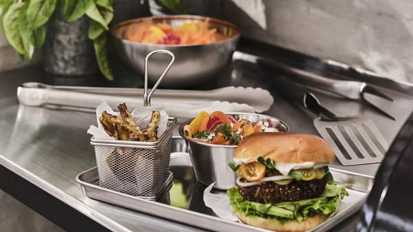 Grillzangen und Burger, Pommes und Grillgemüse in GRILLTIDER Grillzubehör auf einer Edelstahlarbeitsplatte.