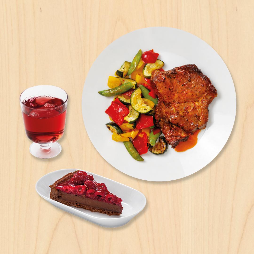 Grilovaná krkovice s grilovanou zeleninou na bílém talíři. K tomu bezedná sklenička s nápojem a čokoládovomalinová porce dortu na oválném talířku.