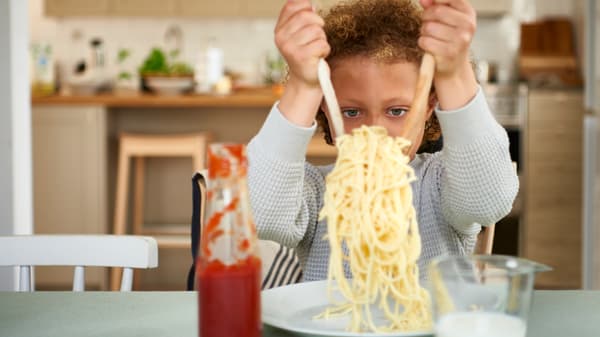 Gripping two wooden utensils, a child in a gray sweater places cooked spaghetti on to a white IKEA 365+ plate.