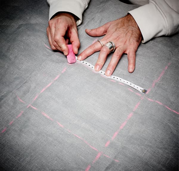 Gros plan de mains tenant une craie de tailleur et traçant un motif en treillis sur du tissu gris.