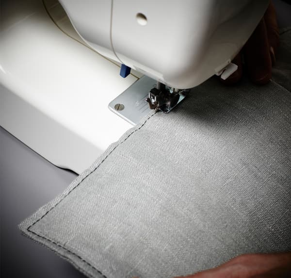 Gros plan d’une machine à coudre IKEA et des mains d’une personne, montrant comment coudre un ourlet sur du tissu gris.