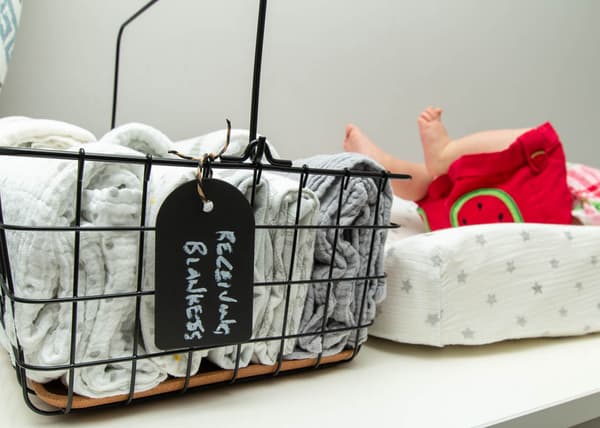 Gros plan sur la corbeille en fil avec poignée PLEJA contenant des couvertures à langer. Un bébé bouge ses pieds en arrière-plan.