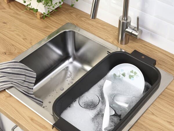 Gros plan sur un évier en inox avec sur la partie droite une bassine noire remplie d'eau mousseuse avec de la vaisselle.