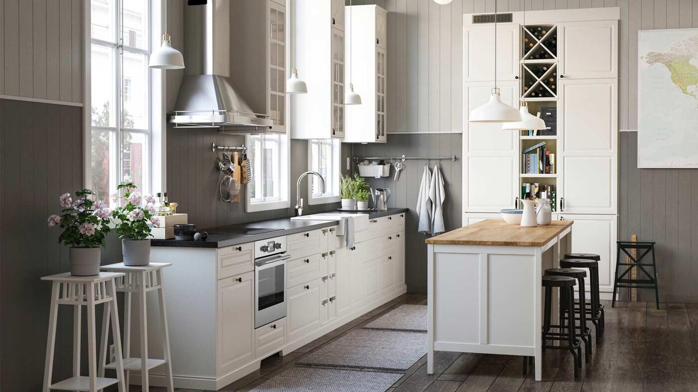 Große IKEA Küchenlösung mit weißen Fronten und weißen Ober- und Unterschränken und einer Kücheninsel. DIe Kücheninsel mit einer Holz Arbeitsplatte und der rest der Küche eine schwarze Arbeitsplatte. Nebenbei stehen 2 Stehplanzentöpfe mit Blumen.