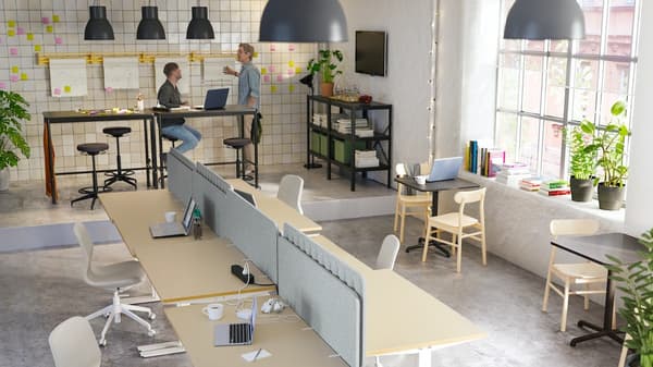 Großes offenes Büro mit zwei jungen Männern, die an hohen Tischen arbeiten, und einer Frau, die auf einem weißen Stuhl an einem Schreibtisch sitzt und E-Mails schreibt.