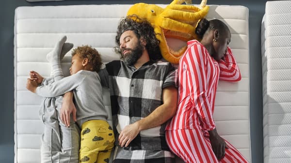 Group of four people lying closely together on white mattresses, wearing colorful pajamas, with a yellow plush toy nearby.