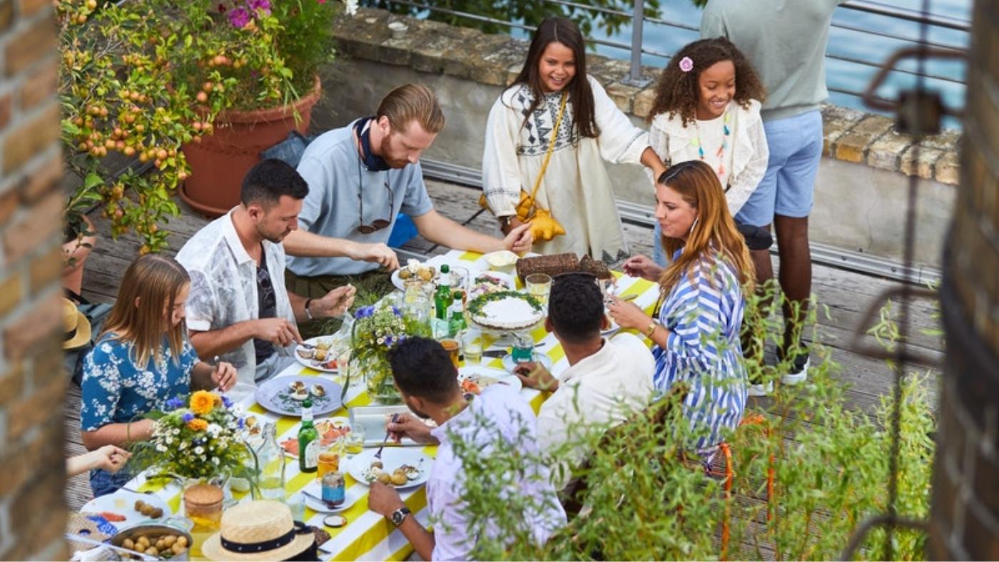 Group of people dressed in white and blue sitting around a table next to a river canal. Table is decorated with flowers and a yellow table cloth.