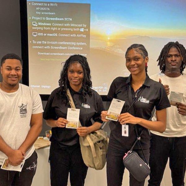 Group of people standing in a classroom holding papers, with a presentation screen displayed behind them.