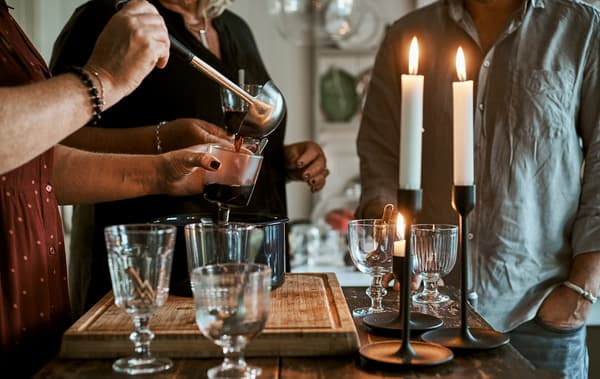 Grupo de persoas ao redor dunha illa de cociña con vasos, candeas acesas e unha pota con ponche quente. Unha muller sirve a bebida nun vaso cun cullerón.