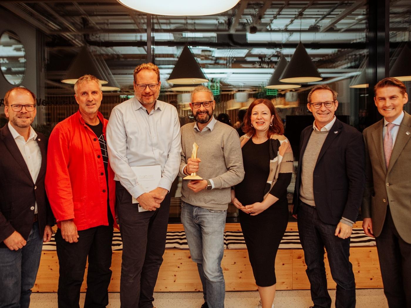 Gruppe von sieben Personen bei einer Auszeichnung in einem modernen IKEA-Umfeld, mittig ein Mann mit Trophäe 