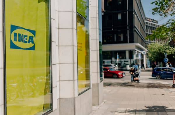 gult skynke med IKEA-logga syns innanför ett fönster som vätter ut mot en gata. I bakgrunden syns man med barnvagn och en röd bil