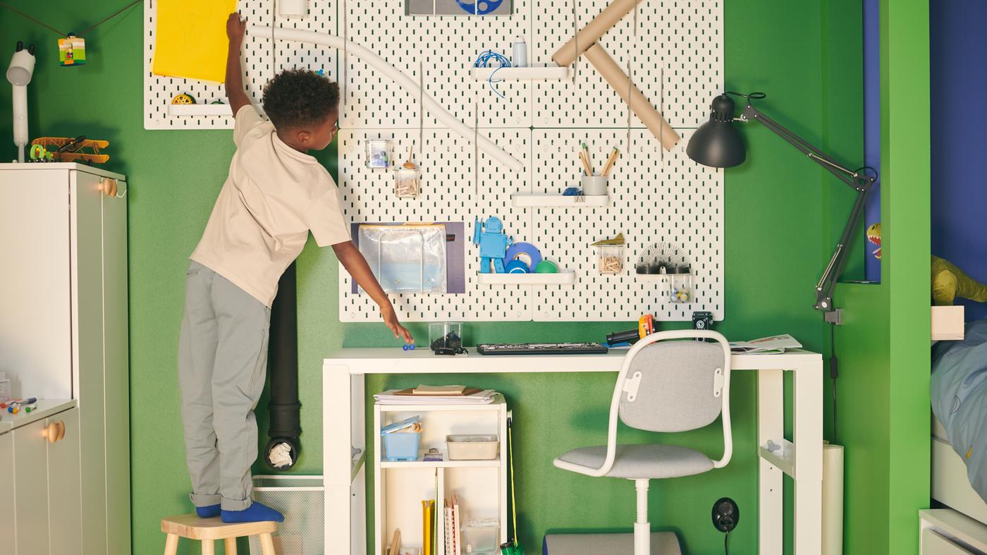 グリーン＆ブルー系の部屋で、スツールの上に立っている男の子が、ホワイトのPÅHL/ポール デスクの上にSKÅDIS/スコーディス ペグボードコンビネーションを掛けている。