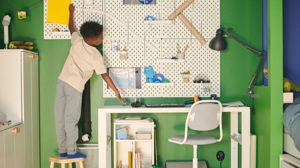 グリーン＆ブルー系の部屋で、スツールの上に立っている男の子が、ホワイトのPÅHL/ポール デスクの上にSKÅDIS/スコーディス ペグボードコンビネーションを掛けている。