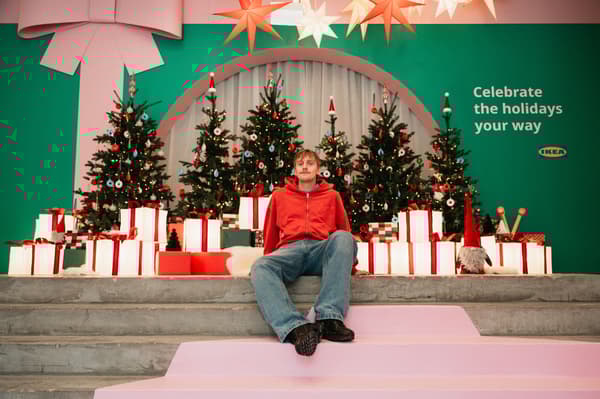 Gustaf Westman sitting on concrete steps against a Christmas-sy backdrop of IKEA Christmas trees, red-and-white wrapped gifts