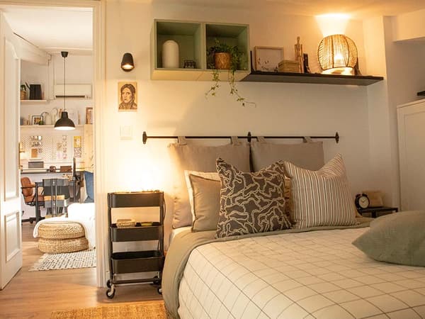Habitació amb un llit, coixins, una tauleta de nit, prestatgeries amb un llum, plantes, fotos i una catifa.