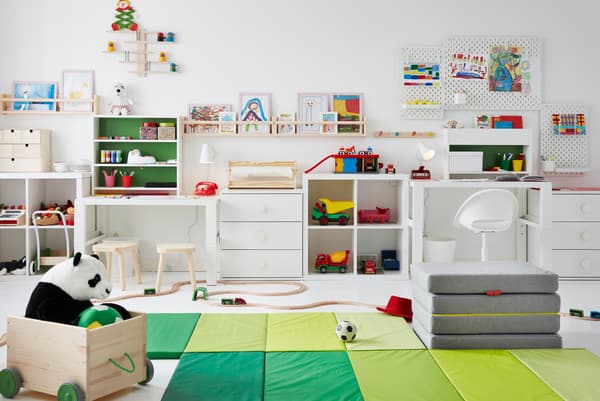Habitació infantil amb joguines a terra, escriptoris als costats i dibuixos emmarcats a sobre de lleixes i taulers perforats SKÅDIS a les parets.