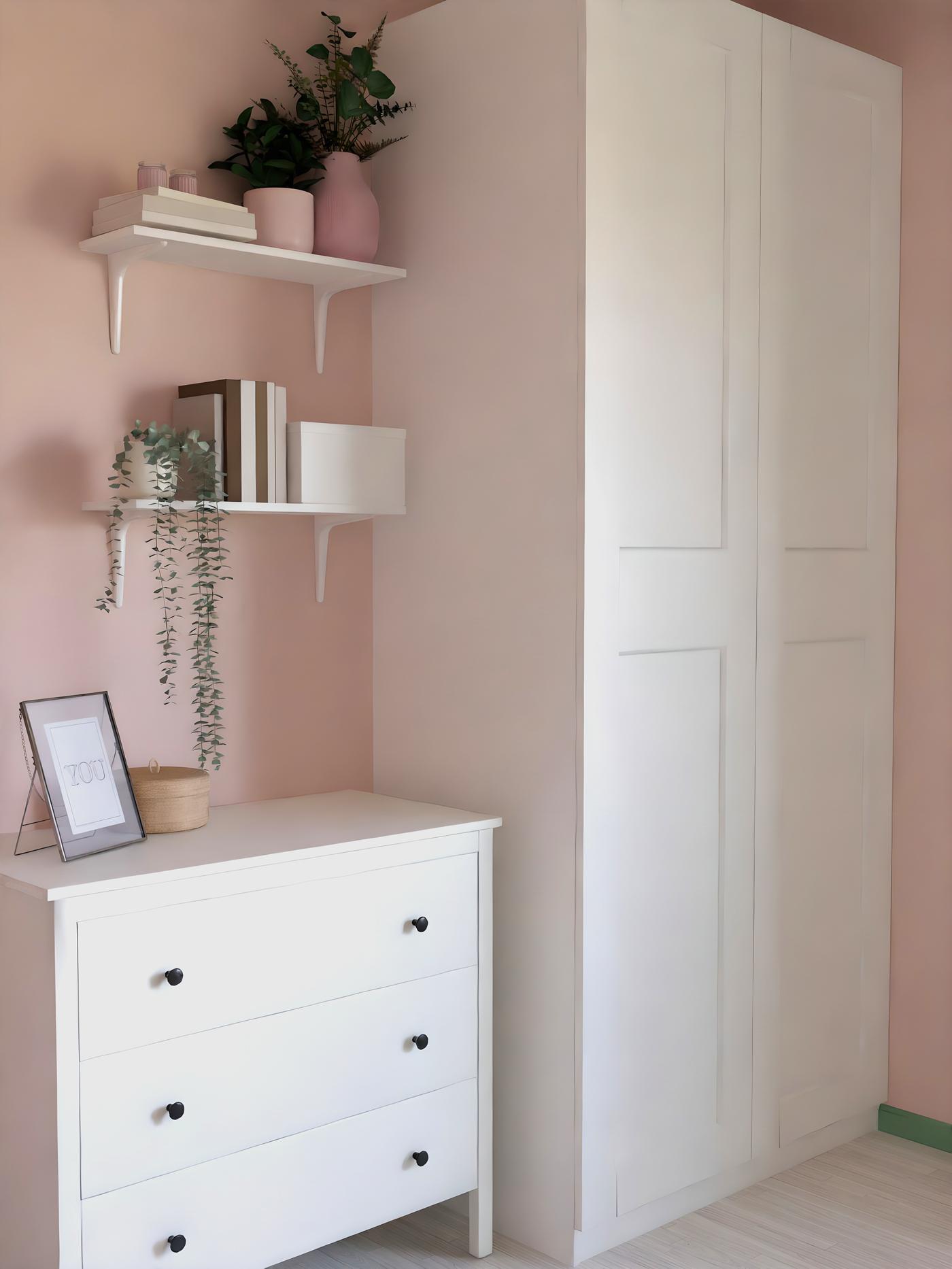 Habitació pintada de rosa amb un armari blanc ampli, una còmoda amb pomos negres i prestatgeries de paret amb llibres i flors.