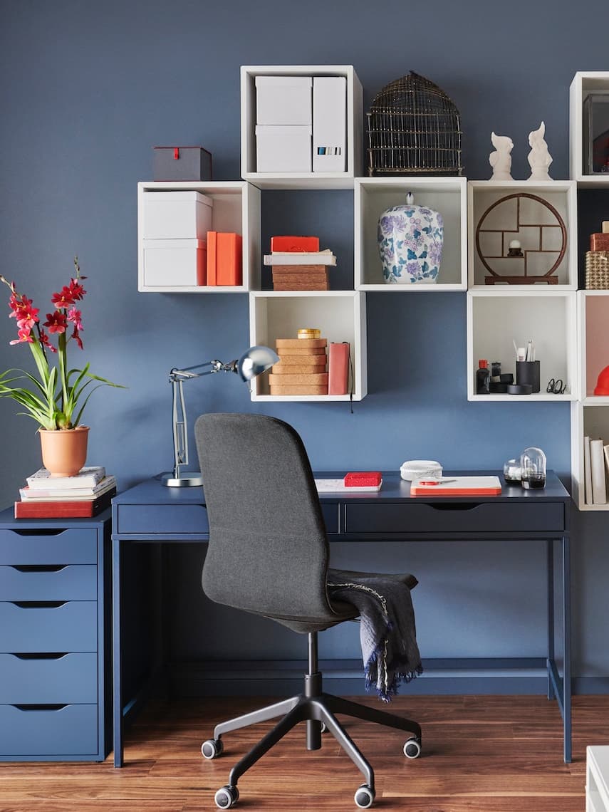 Habitación con pared de color azul con un escritorio azul y repisas de color blanco colgadas en la pared, silla color negro frente al escritorio.