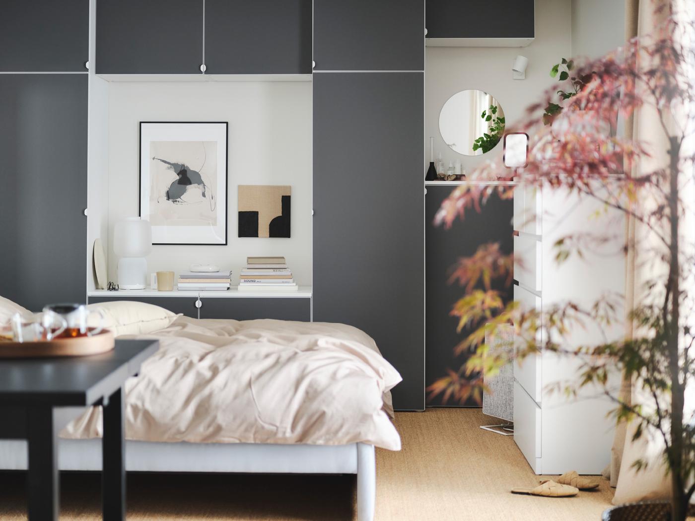 Habitación con una cama con ropa de cama beige, una combinación de armarios grises de fondo, una planta y una mesa negra en primer plano.
