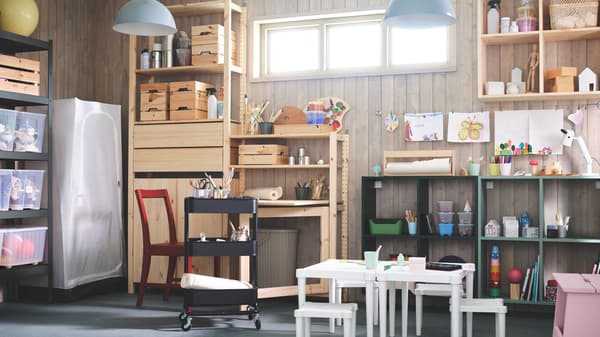 Habitación de un hogar destinado al almacenaje y manualidades en el que aparecen estanterías de madera y metal con cajas, juguetes y utensilios de pintura. Un carrito negro con manualidades y tijeras, mesas y sillas blancas pequeñas y una silla roja. Un armario de tela cerrado.