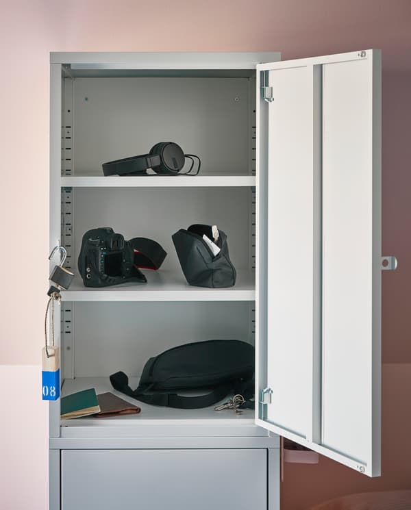 HÄLLAN storage cabinet with the door open showing headphones, camera and other travel belongings on three different shelves.
