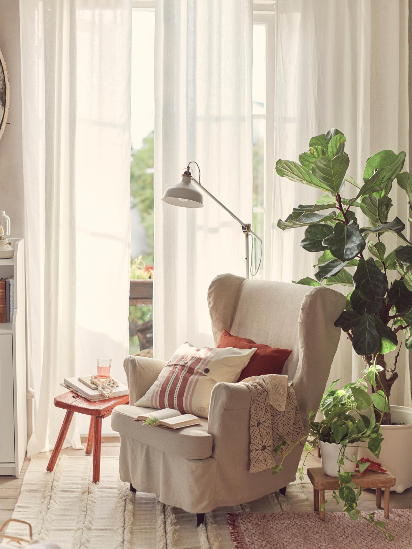 Hagyományos stílusú STRANDMON fotel natúr színű vászon védőhuzattal, párnákkal és takaróval, a cserepes növények társaságában.