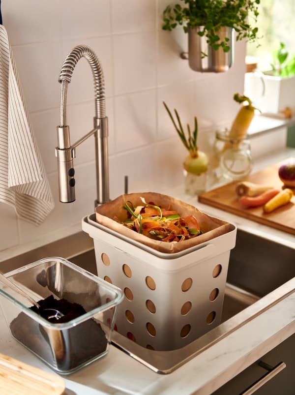 ถุงกระดาษที่เต็มไปด้วยเศษอาหารใส่อยู่ในถังขยะออร์แกนิก HÅLLBAR/ฮลล์บาร์ ที่วางอยู่ในซิงก์ครัว มีโหลใส่กากกาแฟอยู่ข้าง ๆ