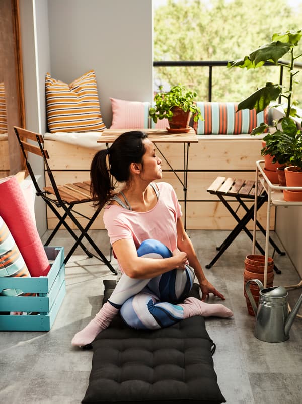 ผู้หญิงในท่าโยคะนั่งบนเบาะรองนั่งและเบาะพิงหลัง HÅLLÖ/โฮลเออ ที่ปูอยู่บนพื้นระเบียงที่มีแสงแดดส่อง