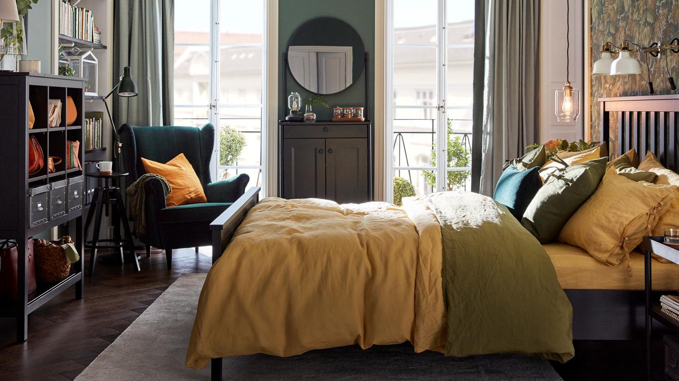 Hálószoba HEMNES ággyal, rajta színes ágynemű, egy zöld STRANDMON szék az egyik sarokban és két erkélyajtó.