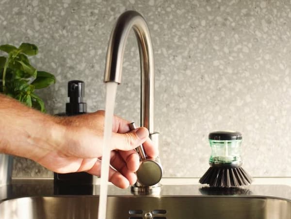 Hand adjusts the water flow of a GLYPEN kitchen tap in stainless steel next to a TÅRTSMET dish-washing brush.