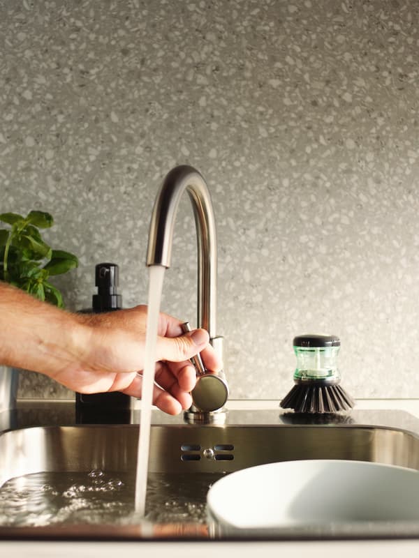 Hand adjusts the water flow of a GLYPEN kitchen tap in stainless steel next to a TÅRTSMET dish-washing brush.