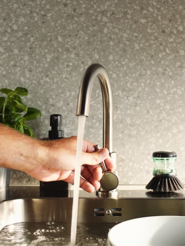 Hand adjusts the water flow of a GLYPEN kitchen tap in stainless steel next to a TÅRTSMET dish-washing brush.