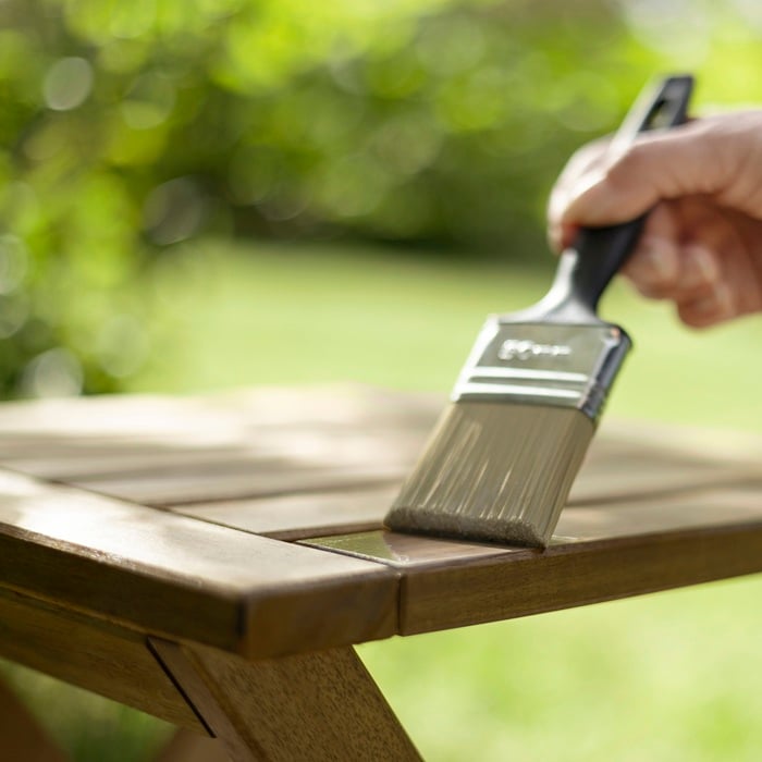 Hand applying wood stain to an outdoor wooden table with a paintbrush.