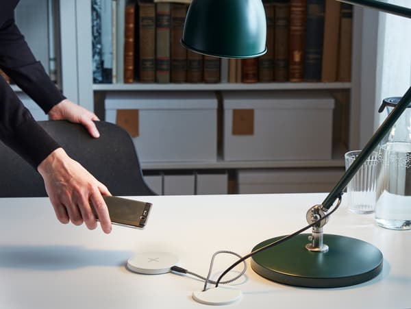 Hand holding a phone over a desktop where the cables of a lamp and a wireless phone charger disappear into the desk.