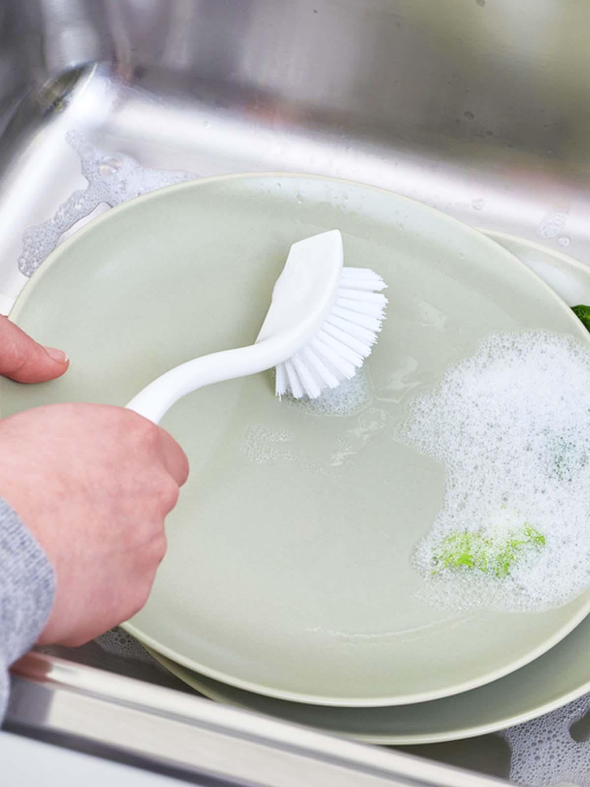 Hand holding ANTAGEN dish-washing brush, white, scrubbing green dishes in the kitchen sink.