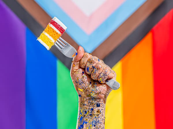 hand holding fork with Rainbow cake, Pride Progress flag in background