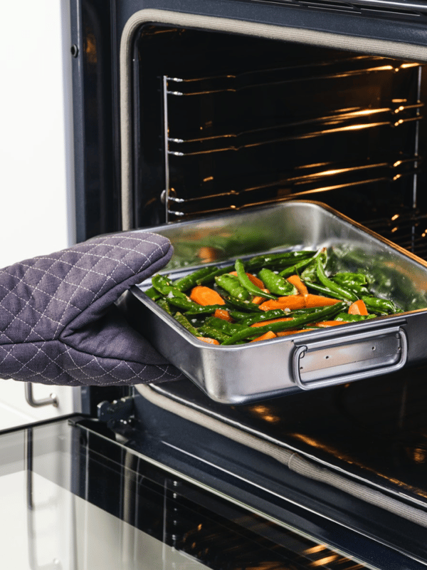 Hand met ovenwant zet een KONCIS braadslede met wortels en andere groenten in de oven.