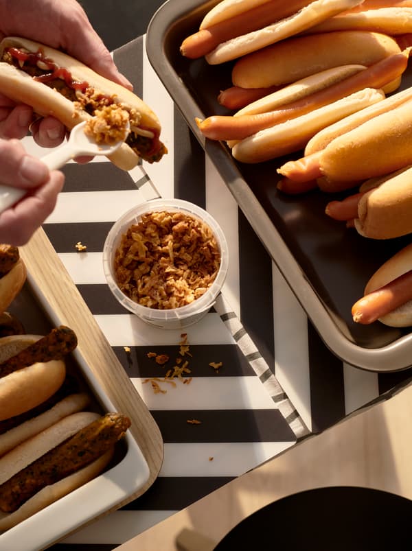 Handen leggen wat ROSTAD LÖK gebakken ui op een KORVMOJ vegetarische hotdog. Op een schotel liggen vegetarische en gewone worsten tussen broodjes.