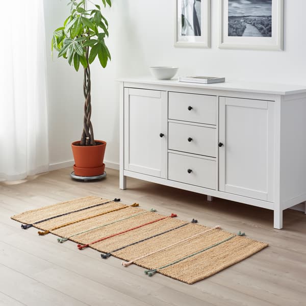 Handgemaakt jute vloerkleed met gekleurde strepen voor een witte ladekast met een plant in de hoek.