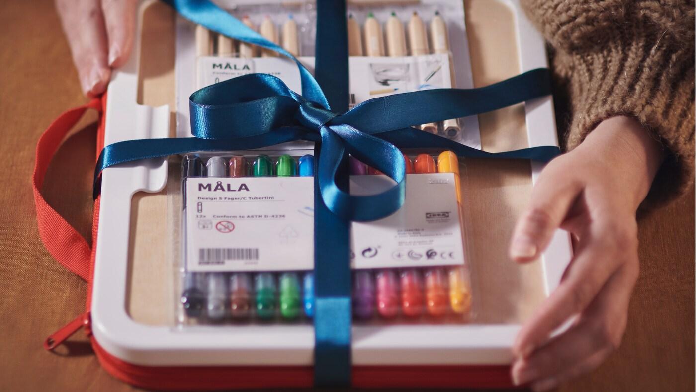 Hands holding a gift box with IKEA MÅLA markers and colored pencils, wrapped with a blue ribbon and bow.