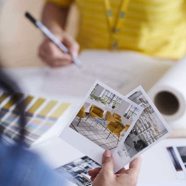 Hands holding printed interior design images over a desk with colour swatches, drawings, and design materials, showing a planning or design process.