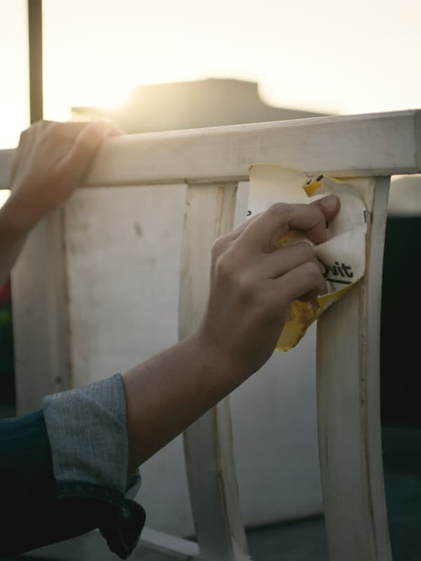 Hands holding sandpaper and sanding a white-painted wooden chair outside with sun glare.