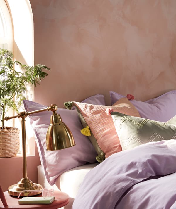Haphazardly made, sunlit bed in which cushions with MOAKAJSA and KLARAFINA covers lie among pillows and bed linen.